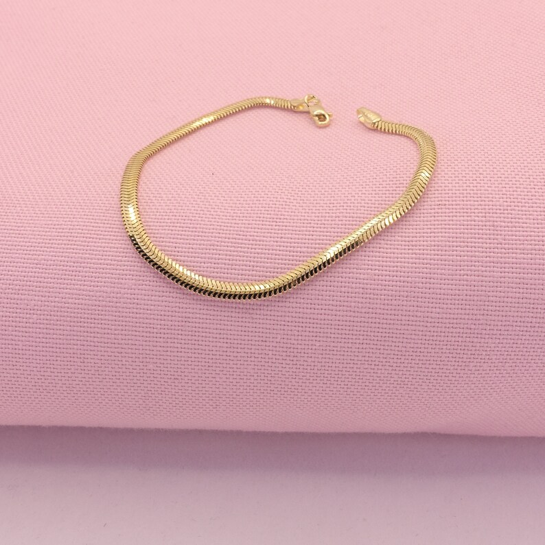 May include: A gold snake chain bracelet on a pink background.
