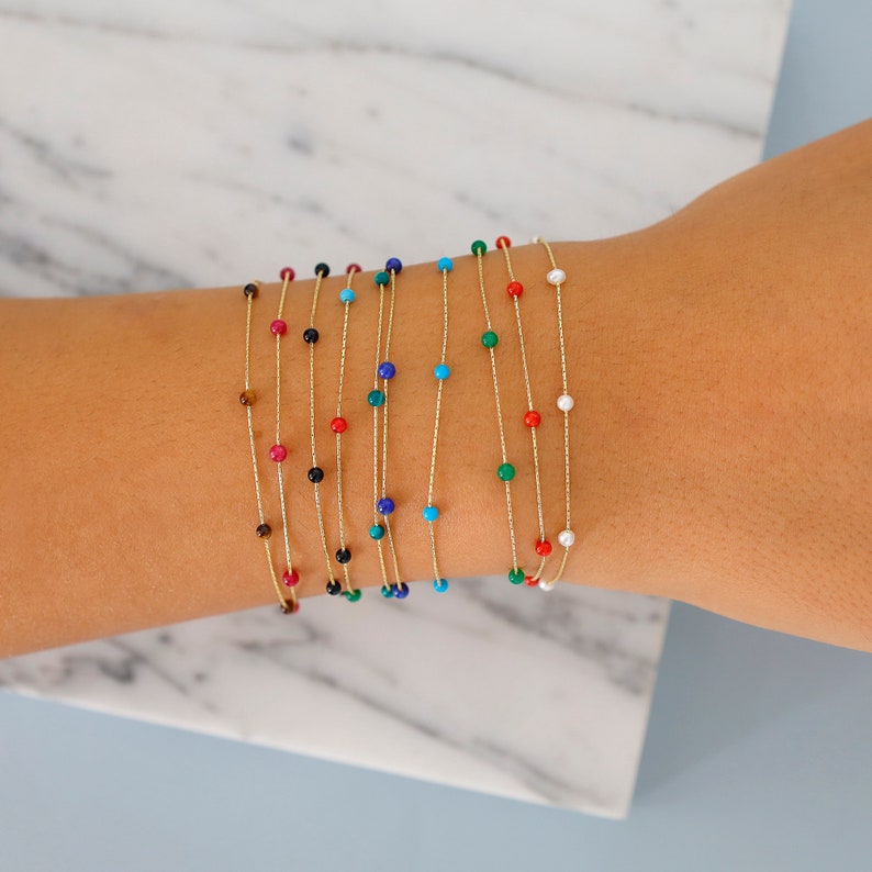 May include: A set of delicate gold chain bracelets with small colourful beads. The bracelets are layered on a person's wrist, showcasing a variety of colours, including red, blue, green, black, and white.