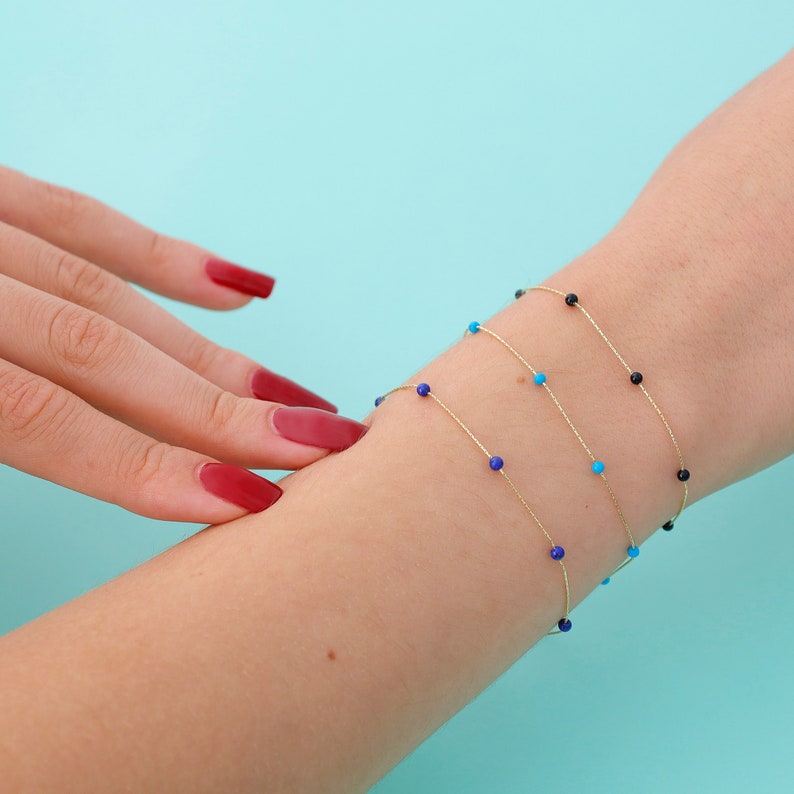 May include: Three delicate gold chain bracelets with small gemstone beads. The bracelets feature blue, turquoise, and black beads.