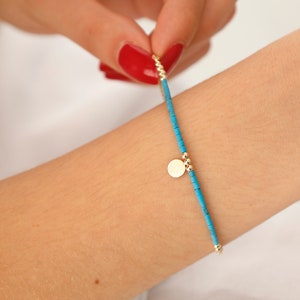 May include: A turquoise beaded bracelet with a gold charm. The bracelet is on a person's wrist.