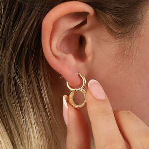 Puede incluir: Pendientes de aro dorados con un diseño circular texturizado. Los pendientes se llevan en la oreja de una persona.