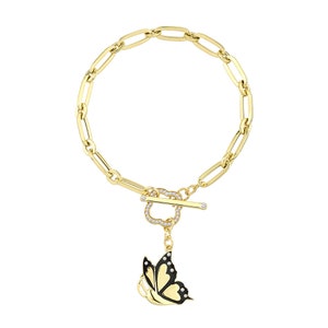 K&ouml;nnte beinhalten: Ein goldenes Kettenarmband mit einem Toggle-Verschluss in Herzform, der mit klaren Steinen besetzt ist. An dem Verschluss h&auml;ngt ein Anh&auml;nger in Form eines schwarzen Schmetterlings mit wei&szlig;en Akzenten.