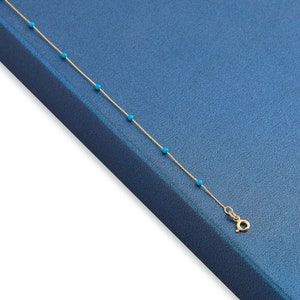 May include: A gold chain necklace with small turquoise beads. The necklace is on a blue background.