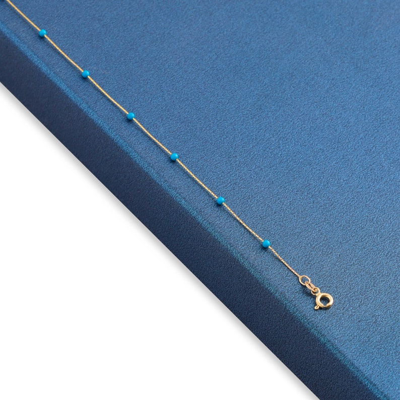 May include: A gold chain necklace with small turquoise beads. The necklace is on a blue background.