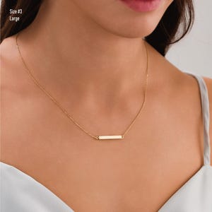 May include: A gold chain necklace with a thin, horizontal bar pendant. The necklace is worn by a person with a light blue top.