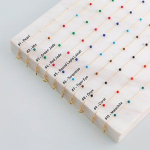 May include: Ten gold chains with different coloured beads, including pearl, turquoise, red jade, lapis lazuli, tiger eye, onyx, coral, and malachite. Each chain has a small gold charm attached.