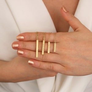 May include: A collection of gold-colored bar pendant necklaces of varying lengths, each suspended from a delicate chain. The bars are engraved with text and symbols. The necklaces are displayed on a person's hand, with a white background.