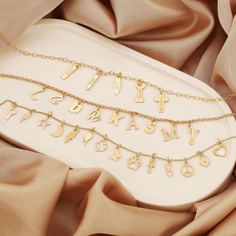 May include: Gold chain necklace with multiple charms, including letters, a cross, a paw print, a feather, a butterfly, a crescent moon, a star, a key, a lightning bolt, a clover, an infinity symbol, a peace sign, and a heart. The charms are attached to the chain with small gold rings.