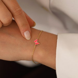 May include: A delicate gold chain bracelet with a pink butterfly charm. The bracelet is made of two thin chains and features a small, detailed butterfly pendant.