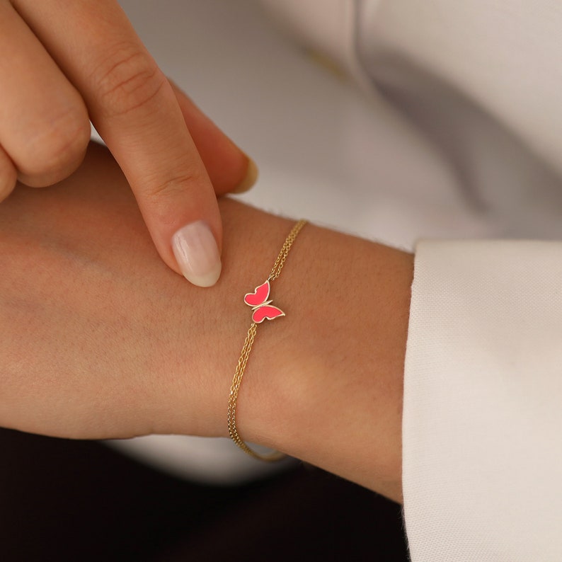 May include: A delicate gold chain bracelet with a pink butterfly charm. The bracelet is made of two thin chains and features a small, detailed butterfly pendant.