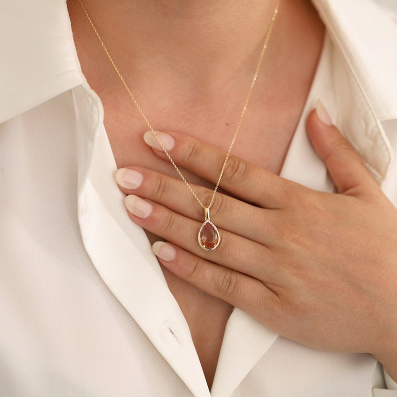 Pu&ograve; includere: Una collana con catena d'oro con un ciondolo a forma di pera con una pietra preziosa marrone circondata da pietre preziose trasparenti pi&ugrave; piccole.