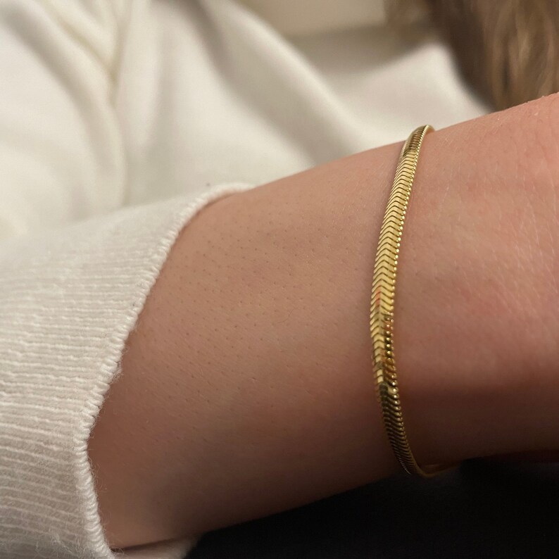 May include: A gold snake chain bracelet on a person's wrist.