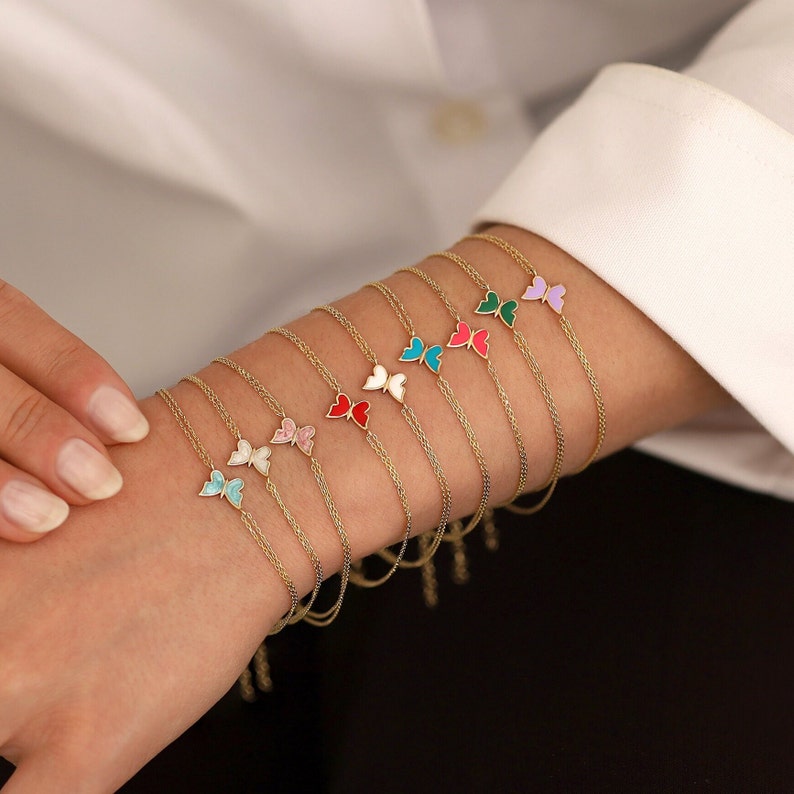 May include: A set of delicate gold chain bracelets with butterfly charms. Each butterfly charm is made of gold and has a different colored enamel coating, including pink, blue, green, red, and purple.