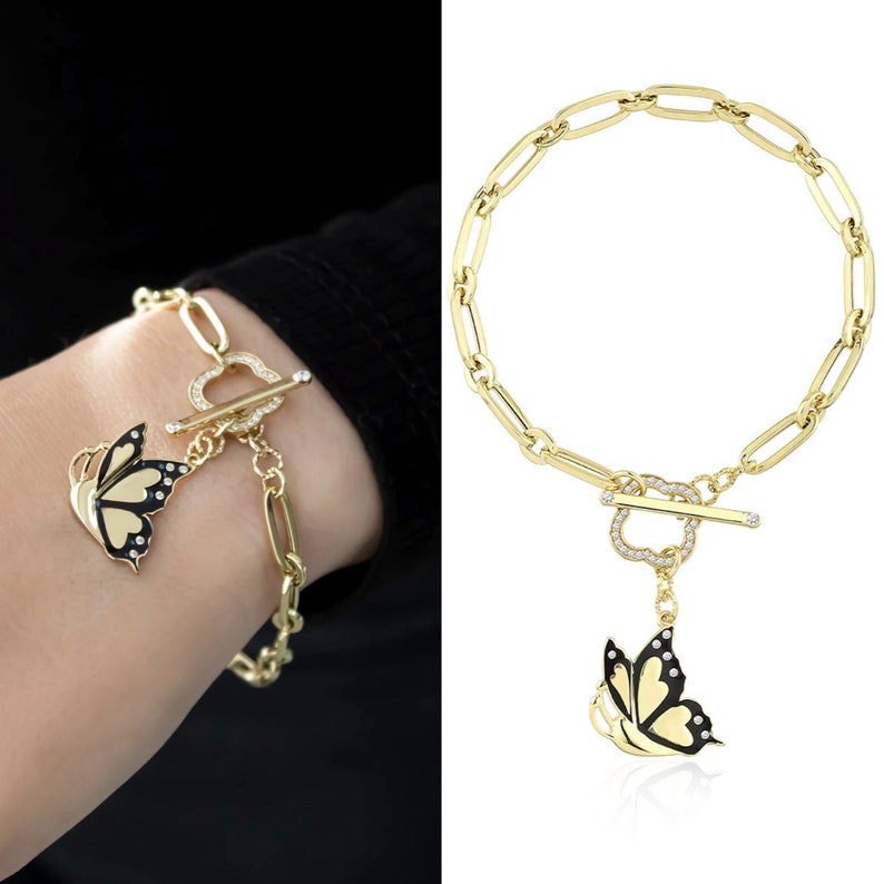 K&ouml;nnte beinhalten: Ein goldenes Kettenarmband mit einem Schmetterlingsanh&auml;nger. Der Schmetterling ist schwarz mit wei&szlig;en Akzenten und hat klare Strasssteine auf seinen Fl&uuml;geln. Das Armband hat einen Toggle-Verschluss.