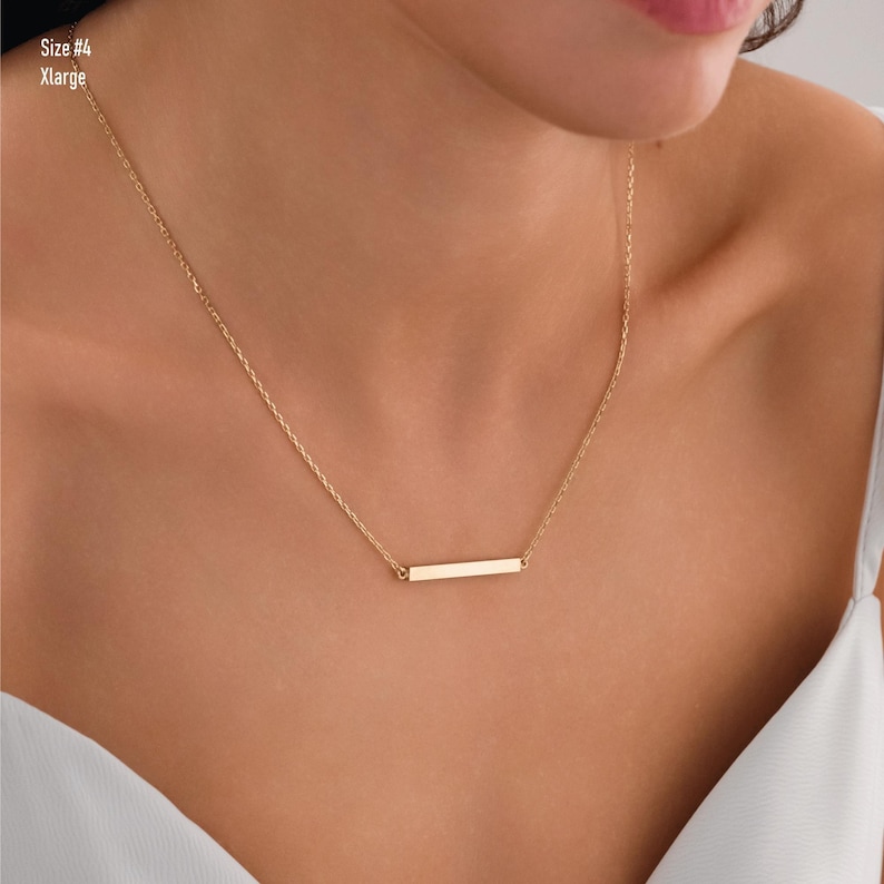 May include: A gold chain necklace with a thin, horizontal bar pendant. The necklace is worn by a person with a light complexion.