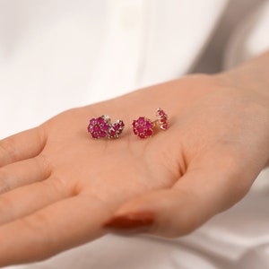 May include: A pair of small, silver stud earrings with a cluster of red ruby stones. The earrings are on a hand, with the stones facing the camera.