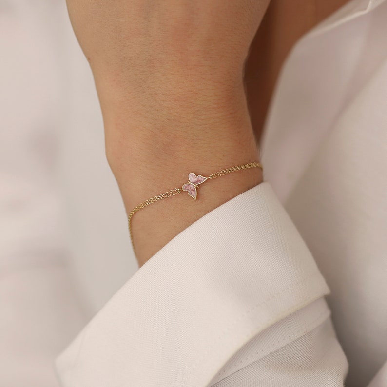 May include: A delicate gold chain bracelet with two pink butterfly charms. The bracelet is on a person's wrist.