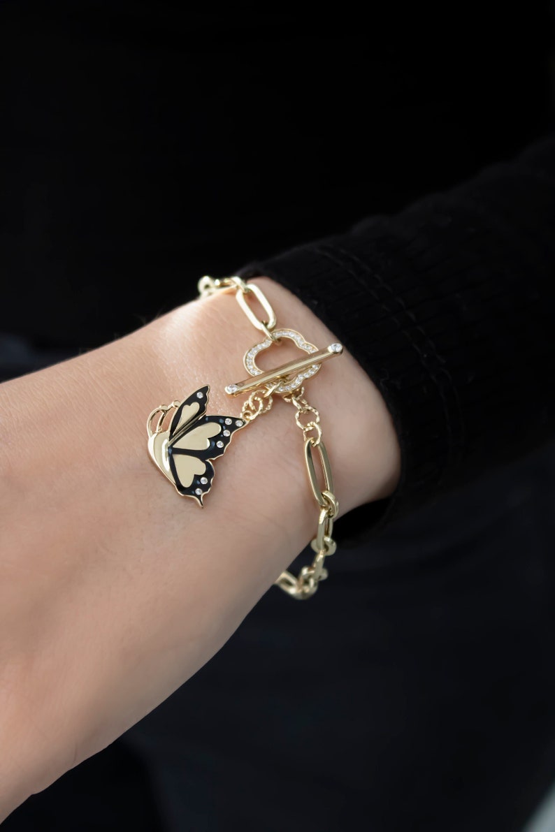 K&ouml;nnte beinhalten: Ein goldenes Kettenarmband mit einem Schmetterlingsanh&auml;nger. Das Armband hat einen herzf&ouml;rmigen Verschluss mit klaren Strasssteinen. Der Schmetterlingsanh&auml;nger ist schwarz und gold mit klaren Strasssteinen.