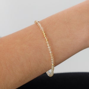 May include: A delicate bracelet made with small, clear beads and gold accents. The bracelet is shown on a person's wrist.