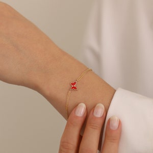 May include: A delicate gold chain bracelet with a red enamel butterfly charm. The bracelet is shown on a person's wrist.