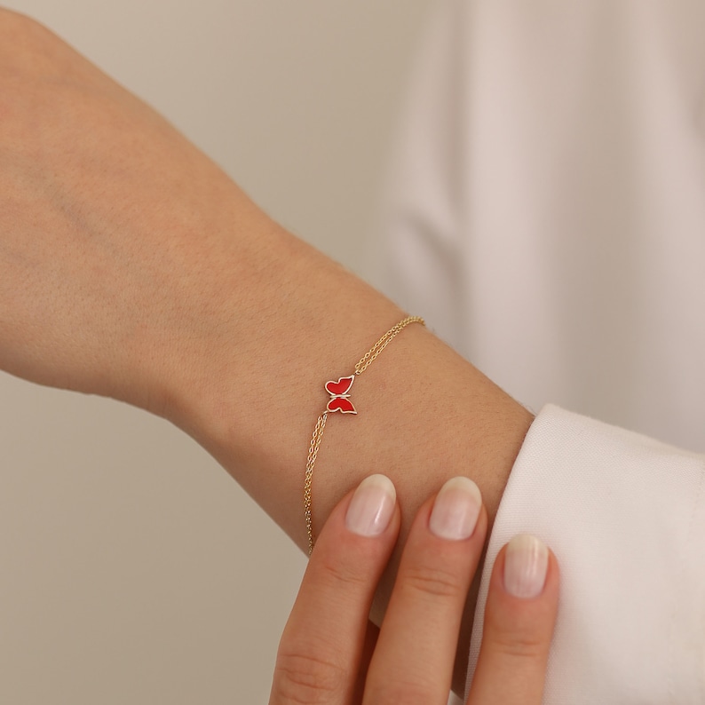 May include: A delicate gold chain bracelet with a red enamel butterfly charm. The bracelet is shown on a person's wrist.