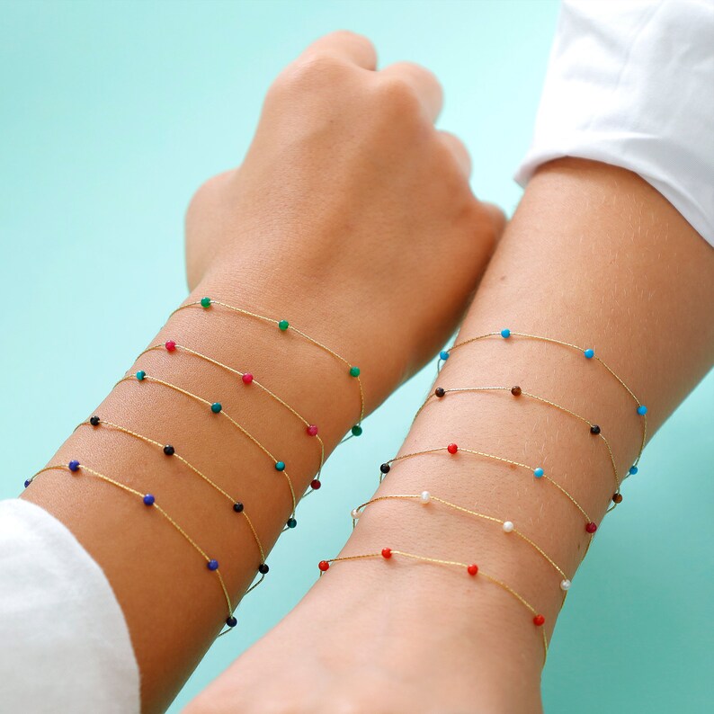 May include: A set of delicate gold bracelets with tiny colorful beads. Each bracelet features a different color bead, including turquoise, red, green, blue, and white.