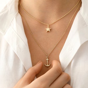 May include: Two gold necklaces with a star and anchor pendant. The star pendant is on a shorter chain and the anchor pendant is on a longer chain.