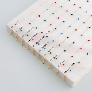 May include: Ten gold chains with different colored beads, including pearl, turquoise, red jade, lapis lazuli, tiger eye, onyx, coral, and malachite. Each chain has a small gold charm attached.