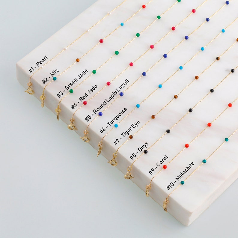 May include: Ten gold chains with different colored beads, including pearl, turquoise, red jade, lapis lazuli, tiger eye, onyx, coral, and malachite. Each chain has a small gold charm attached.