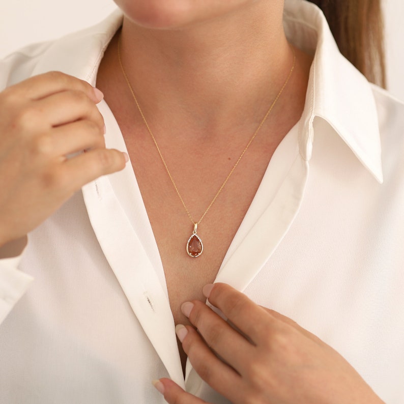 Pu&ograve; includere: Una collana con catena d'oro con un ciondolo a forma di lacrima che presenta una pietra preziosa marrone circondata da un alone di piccole pietre preziose trasparenti.