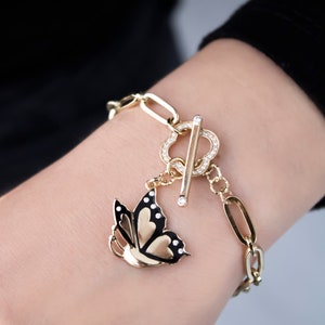 K&ouml;nnte beinhalten: Ein goldenes Kettenarmband mit einem Schmetterlingsanh&auml;nger. Der Schmetterling ist schwarz mit goldenen Akzenten und hat klare Strasssteine. Das Armband hat einen Schiebeverschluss mit klaren Strasssteinen.