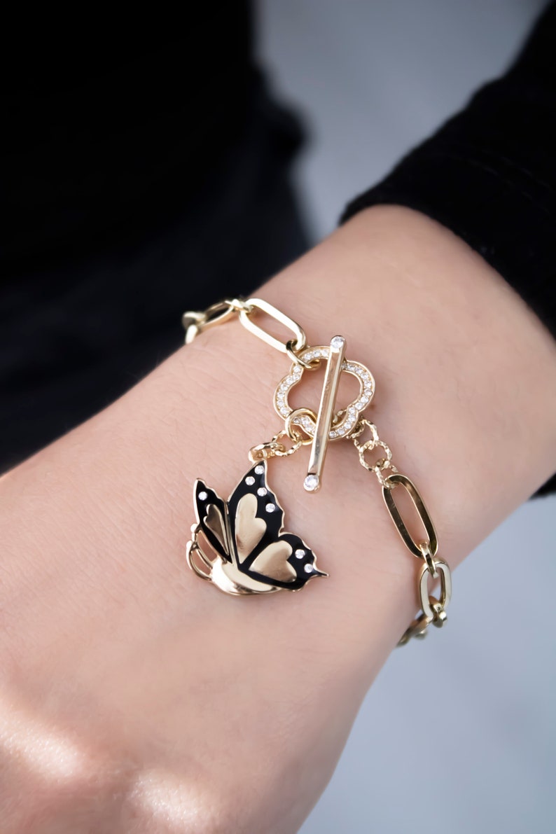 K&ouml;nnte beinhalten: Ein goldenes Kettenarmband mit einem Schmetterlingsanh&auml;nger. Der Schmetterling ist schwarz mit goldenen Akzenten und hat klare Strasssteine. Das Armband hat einen Schiebeverschluss mit klaren Strasssteinen.