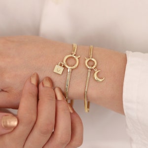 May include: Two gold chain bracelets with a padlock charm and a crescent moon charm. The bracelets are on a person's wrist.