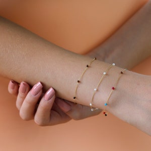 May include: Three delicate gold chain bracelets with small beads. The top bracelet has brown beads, the middle bracelet has white beads, and the bottom bracelet has red and blue beads.