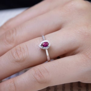 May include: A silver ring featuring a pear-shaped, deep red gemstone surrounded by a halo of small, clear stones. The ring is set on a thin, polished band, showcasing a classic and elegant design.