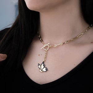 K&ouml;nnte beinhalten: Eine goldene Kettenkette mit einem Schmetterlingsanh&auml;nger. Der Schmetterling ist schwarz mit wei&szlig;en Akzenten und hat einen goldenen Umriss. Die Kette hat einen Toggle-Verschluss.