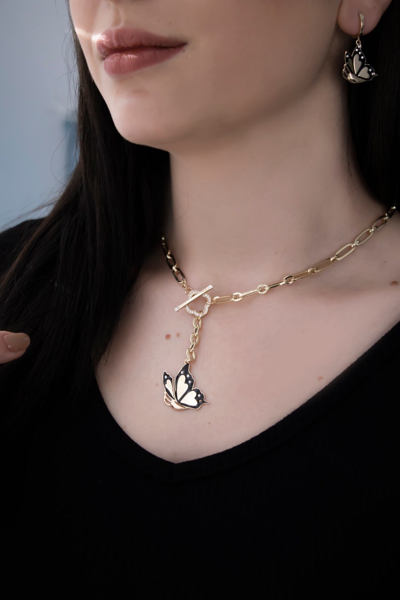 K&ouml;nnte beinhalten: Eine goldene Kettenkette mit einem Schmetterlingsanh&auml;nger. Der Schmetterling ist schwarz mit wei&szlig;en Akzenten und hat einen goldenen Umriss. Die Kette hat einen Toggle-Verschluss.