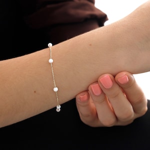 Könnte beinhalten: Ein zartes Armband aus einer goldenen Kette mit kleinen weißen Perlen. Das Armband befindet sich am Handgelenk einer Person.