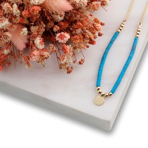 May include: A turquoise blue beaded necklace with a gold pendant and a gold chain. The necklace is on a white marble surface with dried flowers.