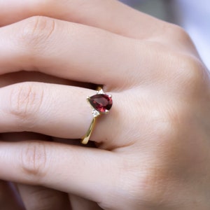 Op de afbeelding: Een gouden ring met een peervormige rode edelsteen en kleinere heldere edelstenen op de band.