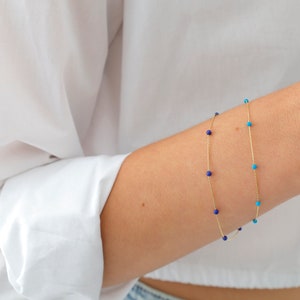 May include: Two delicate gold bracelets with small blue and turquoise beads. The bracelets are layered on a person's wrist.