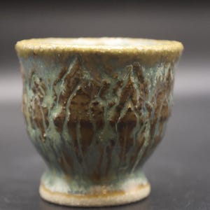 May include: A handmade ceramic cup with a textured, organic design. The cup features a footed base and a flared rim. The exterior is glazed in shades of brown and teal, creating a unique, earthy aesthetic. The cup is suitable for beverages.