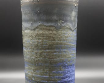 May include: A tall, cylindrical ceramic vase with a textured surface. The vase features a gradient of colors, transitioning from a dark gray rim to olive green and blue bands. The base is a light cream color.