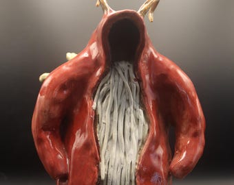 May include: A ceramic sculpture of a hooded figure in a red robe. The figure has a hollow face, white beard-like strands, and golden horns. The figure's feet are light green with brown hooves. The sculpture is on a black background.