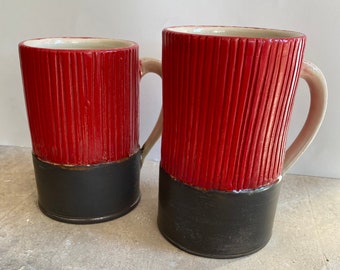 May include: Two ceramic mugs with a red and black design. The mugs have a textured, ribbed surface and a black base.