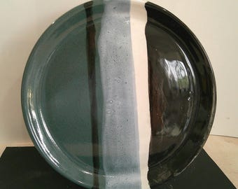 May include: A large, round ceramic plate with a glossy finish. The plate is divided into vertical sections of teal, gray, white, and black. The plate is sitting on a black surface.