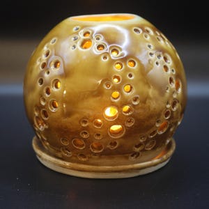 May include: A brown ceramic candle holder with numerous circular openings. The spherical holder sits on a matching base. The interior glows with warm light, creating a cozy ambiance. The surface has a glossy finish.