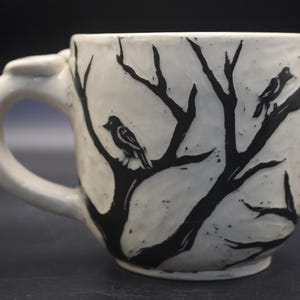 May include: A white ceramic mug featuring a hand-carved black graphic design of bare tree branches and two birds perched on the limbs. The mug has a thick, curved handle and a textured, uneven surface finish throughout the exterior body.