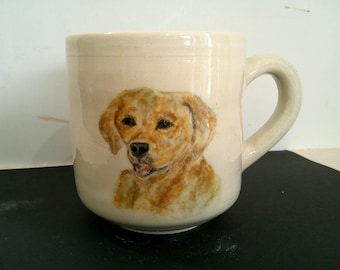 May include: Cream-colored ceramic mug featuring a hand-painted portrait of a golden retriever. The dog's fur is rendered in shades of brown and tan, with a black nose and a pink tongue. The mug has a curved handle.