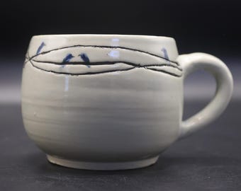 May include: A light gray ceramic mug with a rounded body and a side handle. The exterior features a hand-drawn black line encircling the upper portion, depicting a wire with small blue birds perched upon it, finished with a glossy glaze.
